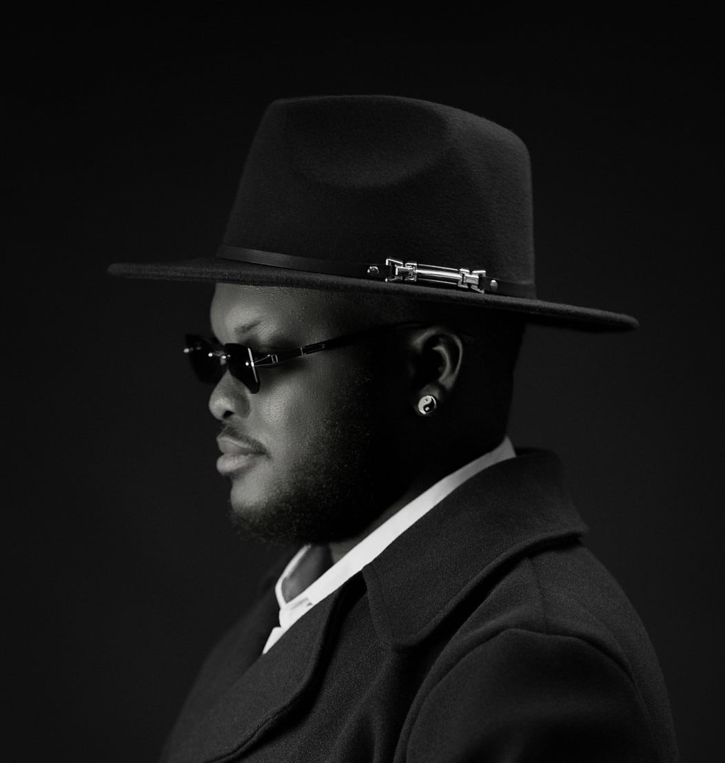 Ayoola Abiola in profile, wearing a fedora and dark coat